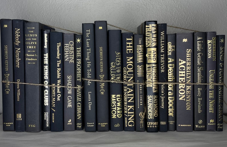 May include: A collection of black hardcover books with gold lettering, arranged on a shelf and bound with twine. Titles include "Nobody Nowhere" and "The Mountain King". The books create a sophisticated, minimalist aesthetic.