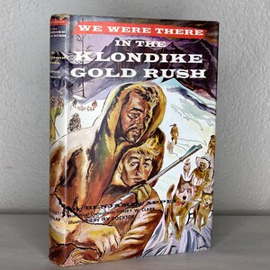 Op de afbeelding: Boekomslagillustratie van twee mannen in winterkleding met een hondensleeteam op de achtergrond. De titel van het boek is "We Were There in the Klondike Gold Rush" door Benjamin Appel.