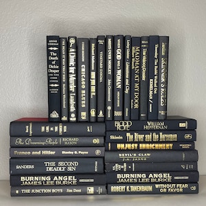 May include: A collection of hardcover books with black covers and gold lettering. The titles and authors are visible, including "Burning Angel" and "The Death of Dickie Draper." The books are stacked neatly, creating a visually appealing arrangement.