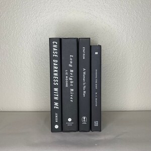 May include: A stack of five hardcover books with black covers. The titles and authors are visible, including "Chase Darkness With Me" by Jensen, "Long Bright River" by Liz Moore, and "A Woman Is No Man". The books are arranged vertically.
