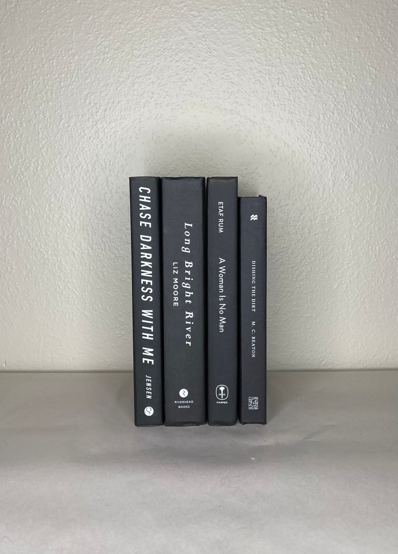 May include: A stack of five hardcover books with black covers. The titles and authors are visible, including "Chase Darkness With Me" by Jensen, "Long Bright River" by Liz Moore, and "A Woman Is No Man". The books are arranged vertically.