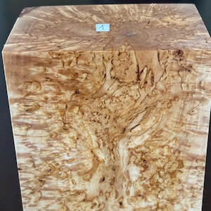 Könnte beinhalten: Ein rechteckiger Block aus hellbraunem Holz mit einer dichten, wirbelnden Maserung auf der gesamten Oberfläche. Ein kleines weißes rechteckiges Etikett mit der in Schwarz aufgedruckten Zahl 1 ist oben mittig auf dem Holzblock angebracht.