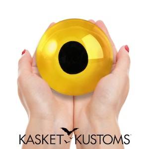 May include: A pair of hands holding a large, golden eyeball with a black pupil. The hands are against a white background.