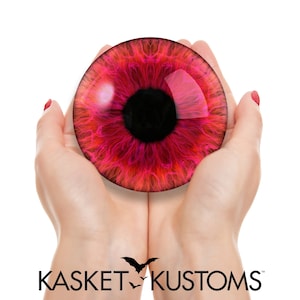 May include: Two hands holding a large, round, red eye with a black pupil. The eye has a pink and orange iris with a glossy finish.