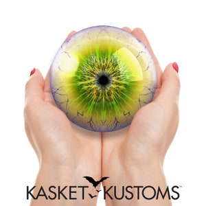 May include: A pair of hands holding a large, round, glass eye with a yellow and green iris and a black pupil. The eye is surrounded by a purple and white vein-like pattern.