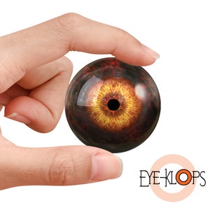 Puede incluir: Primer plano de un ojo de vidrio de aspecto realista con un iris marrón oscuro y una pupila naranja brillante. El ojo está sostenido en una mano, y el fondo es blanco.