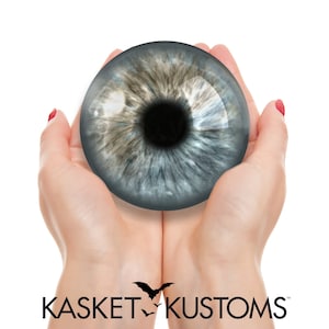 Puede incluir: Un par de manos sosteniendo un gran accesorio de ojo azul realista. El ojo tiene una pupila oscura y un iris marrón claro. Las manos llevan esmalte de uñas rojo.