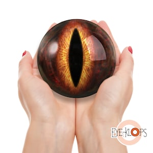 May include: A pair of hands holding a large, round, orange and black eye with a vertical pupil. The eye is glossy and appears to be made of glass.