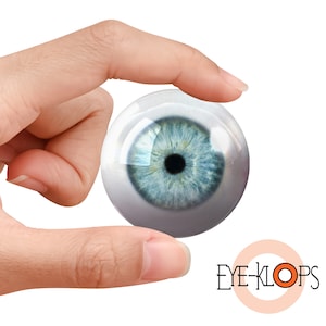 Puede incluir: Primer plano de un ojo azul, con esclerótica blanca y pupila negra, sostenido entre dos dedos. El ojo es un accesorio realista, y la imagen está aislada sobre un fondo blanco. El texto "EYE-KLOPS" es visible en la esquina inferior derecha de la imagen.