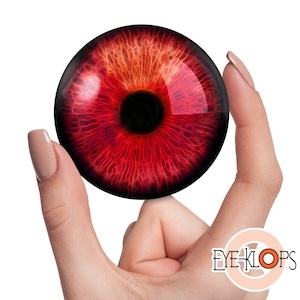 Puede incluir: Primer plano de un accesorio de ojo rojo con pupila negra e iris rojo detallado. El ojo se sostiene entre dos dedos.