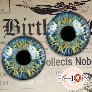 May include: Two blue and yellow realistic eye glass cabochons with black pupils. The cabochons are on a vintage newspaper background with the word "Birth" printed in black.