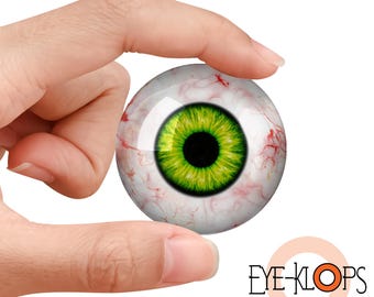 May include: A realistic, glass eye with a vibrant green iris and a black pupil. The eye is held between fingers, showcasing the intricate details of the veins and the clear, glossy surface. The logo "EYE-KLOPS" is visible.