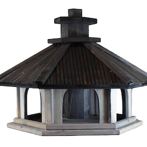 May include: A wooden birdhouse with a pagoda-style roof. The roof is dark brown, and the body is a mix of light and dark gray wood. The birdhouse has an octagonal base and a small, square top.
