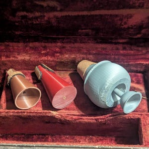 May include: Three musical instrument mutes in a red velvet-lined case. The mutes are copper, red, and gray. The copper mute is cylindrical, the red mute is conical, and the gray mute is bulbous with a small bell.