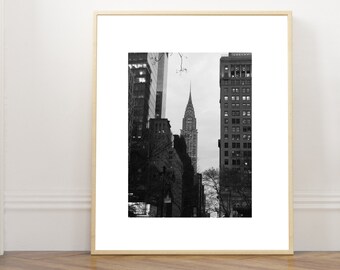 Könnte beinhalten: Ein schwarz-weiß gerahmter Druck mit einer Ansicht des Chrysler Buildings in New York City, gerahmt in einem hellen Holzrahmen mit einem breiten weißen Passepartout, das den Wolkenkratzer zwischen zwei höheren Gebäuden mit kahlen Ästen zeigt.
