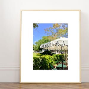 Op de afbeelding: Een ingelijste print met een witte terrasparasol met een geschulpte rand en zwarte afwerking, geplaatst boven een kleine groene tafel en stoelen in een weelderige tuin met een blauwe lucht en groene bomen op de achtergrond.