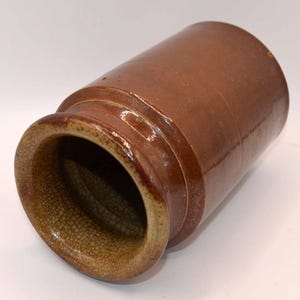 May include: A cylindrical ceramic crock featuring a smooth, dark brown glazed exterior body and a contrasting light tan interior rim with a visible crackle glaze texture. The vessel has a wide, open mouth and a sturdy, thick-walled construction.