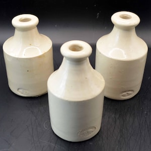 May include: Three off-white ceramic bottles with cylindrical bodies and narrow, rounded necks. Each bottle features a smooth, glazed finish and a small, oval-shaped embossed stamp near the base, showing a consistent design across all three items.
