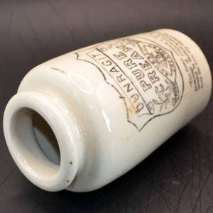 May include: A cylindrical off-white ceramic jar with a wide opening. The exterior features black printed text that reads DUNRAGIT PURE CREAM inside a shield-shaped emblem, along with additional small text printed on the side of the vessel.