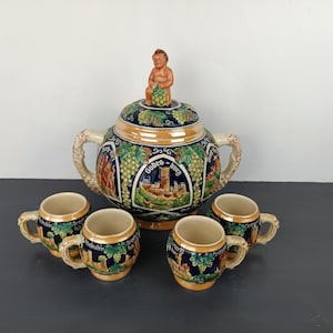Può includere: Una zuppiera in ceramica con un coperchio che presenta una piccola statuina di un uomo che tiene dell'uva. La zuppiera è decorata con un design blu e oro e presenta una scena di un castello con il testo "Godefrid - Burg". La zuppiera è circondata da quattro tazze in ceramica abbinate.