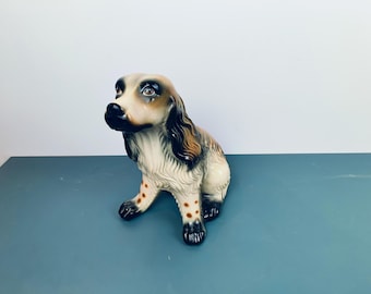 Peut inclure: Une figurine en céramique d'un chien brun et blanc avec des yeux noirs et un nez brun. Le chien est assis, les pattes croisées et la queue enroulée autour de son corps.