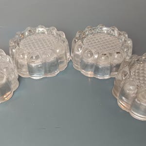Puede incluir: Un juego de cuatro posavasos de vidrio transparente, cada uno con forma circular, un patrón de cuadrícula texturizado en la superficie superior y un diseño de borde festoneado similar a pétalos alrededor del perímetro, con aspecto reflectante.