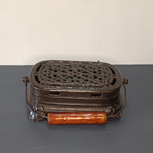 May include: A vintage cast iron stove top warmer with a decorative grill design and a wooden handle. The warmer is black and has a rustic look.