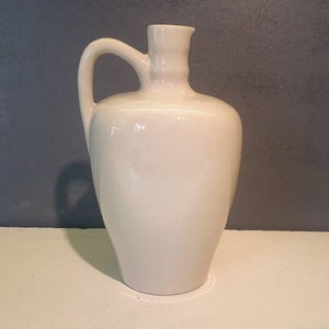 May include: A white ceramic pitcher with a rounded body and a handle. The pitcher has a smooth, glossy finish.