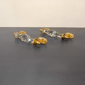 Peut inclure: Deux poignées de porte de placard en verre transparent avec des accents en métal doré. Les poignées sont facettées et présentent un design décoratif.