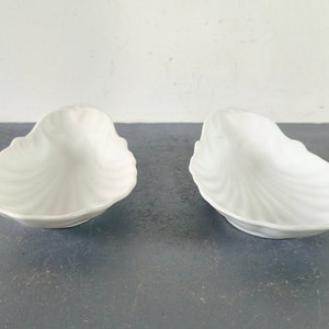 May include: Two white ceramic dishes shaped like seashells. The dishes are smooth and have a glossy finish.
