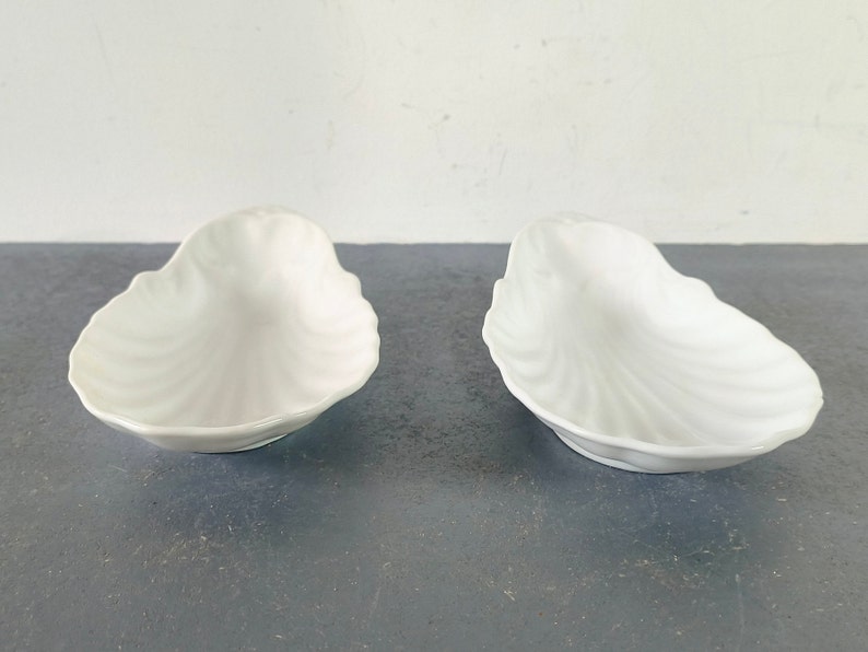 May include: Two white ceramic dishes shaped like seashells. The dishes are smooth and have a glossy finish.