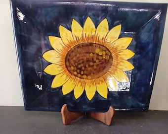 May include: A square ceramic plate with a blue glaze and a hand-painted sunflower design. The sunflower is yellow with a brown center and is set against a dark blue background.