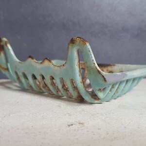 Pu&ograve; includere: Un portasapone vintage in ghisa, di colore azzurro chiaro, con un design traforato decorativo. Il portasapone ha una forma rettangolare e presenta segni di ruggine e usura.