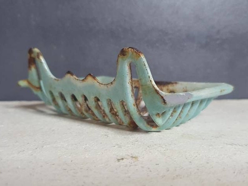 Pu&ograve; includere: Un portasapone vintage in ghisa, di colore azzurro chiaro, con un design traforato decorativo. Il portasapone ha una forma rettangolare e presenta segni di ruggine e usura.