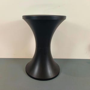 Puede incluir: Un taburete negro mate con una silueta lisa en forma de reloj de arena, que presenta un asiento circular y una base circular a juego. El diseño consta de dos secciones cóncavas simétricas que se unen en una costura central continua.
