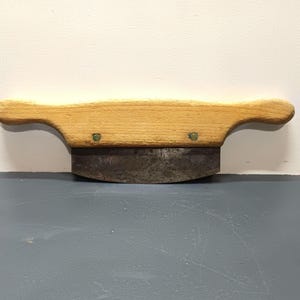 May include: A wooden-handled, metal-bladed scraper tool with two brass rivets. The blade is curved and the handle is a light brown color.
