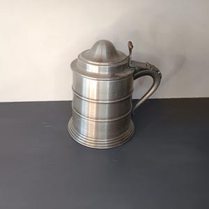 Può includere: Un boccale di birra in peltro argentato con un coperchio incernierato e un manico. Il boccale ha un corpo cilindrico con una parte superiore arrotondata e una base svasata.