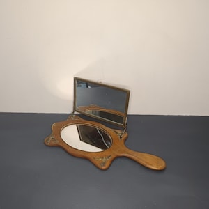 Op de afbeelding: Een houten handspiegel met een gesneden ontwerp en een kleinere spiegel die aan de achterkant is bevestigd. De spiegel heeft een bruine houten handgreep.