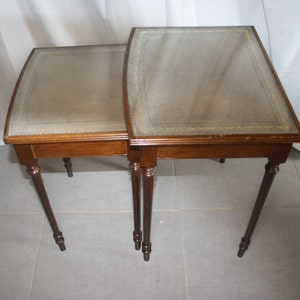 Può includere: Un set di due tavolini a incastro con gambe in legno marrone e piano in pelle marrone.