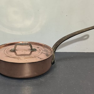 Può includere: Una pentola di rame con coperchio e un lungo manico marrone scuro. La pentola è lucida e ha un design classico.