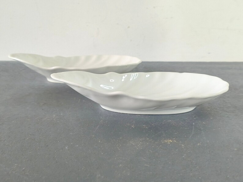 May include: Two white ceramic bowls shaped like seashells. The bowls are smooth and have a glossy finish.