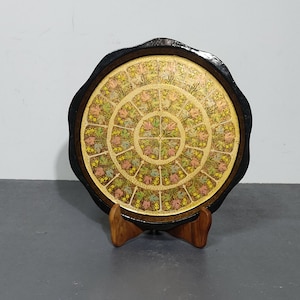 Pode incluir: Uma bandeja redonda decorativa com uma borda preta e um interior dourado. A bandeja é decorada com um padrão floral em tons de rosa, amarelo e verde. A bandeja está em um suporte de madeira.