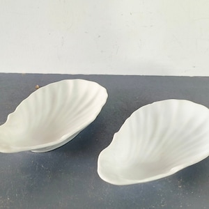 May include: Two white ceramic dishes shaped like seashells. The dishes are smooth and have a glossy finish.