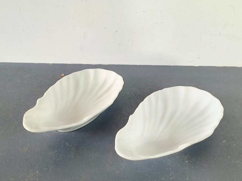 May include: Two white ceramic dishes shaped like seashells. The dishes are smooth and have a glossy finish.