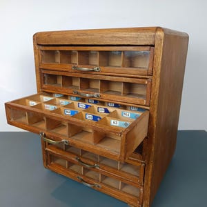 Könnte beinhalten: Ein Holzschrank mit fünf horizontalen Schubladen, die jeweils eine Glasfront und einen Metallgriff haben. Die Schubladen sind in mehrere kleine Fächer unterteilt, wobei im offenen Fach mehrere blau-weiße nummerierte Etiketten zu sehen sind.