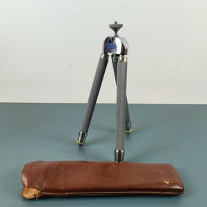Peut inclure: Un petit trépied argenté et gris à trois pieds avec un logo bleu. Il est accompagné d'un étui en cuir marron. Les pieds ont une prise texturée. Le trépied est conçu pour supporter un appareil photo, et mesure environ 20 cm de haut.