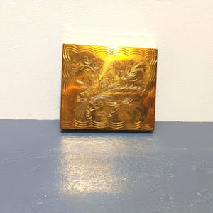 Op de afbeelding: Een gouden metalen compact met een gegraveerd bloemenontwerp op de bovenkant.