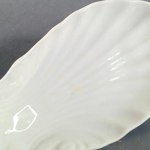May include: White ceramic dish shaped like a seashell. The dish has a smooth, glossy finish and a slightly rippled texture.