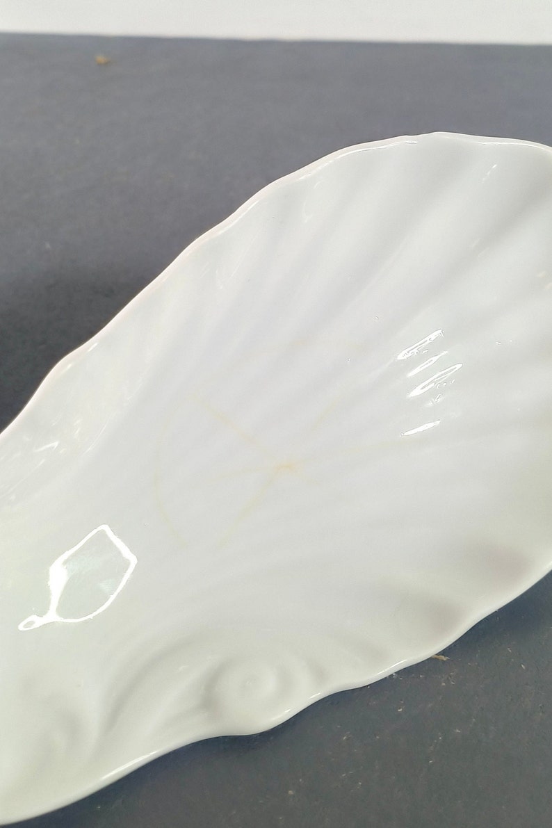 May include: White ceramic dish shaped like a seashell. The dish has a smooth, glossy finish and a slightly rippled texture.