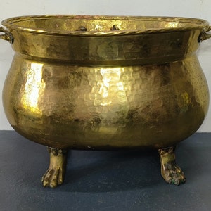 Può includere: Un grande vaso da fiori in ottone martellato con due manici e tre piedi a forma di zampe di animali. Il vaso è di colore dorato e ha una finitura lucida.